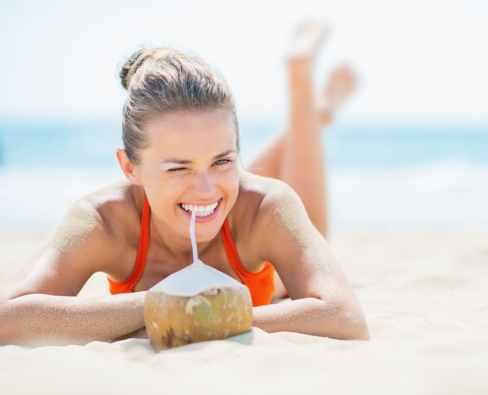 8 Tipps für heiße Tage Beitragsbild: Frau liegt im Bikini mit Kokosnuss am Strand